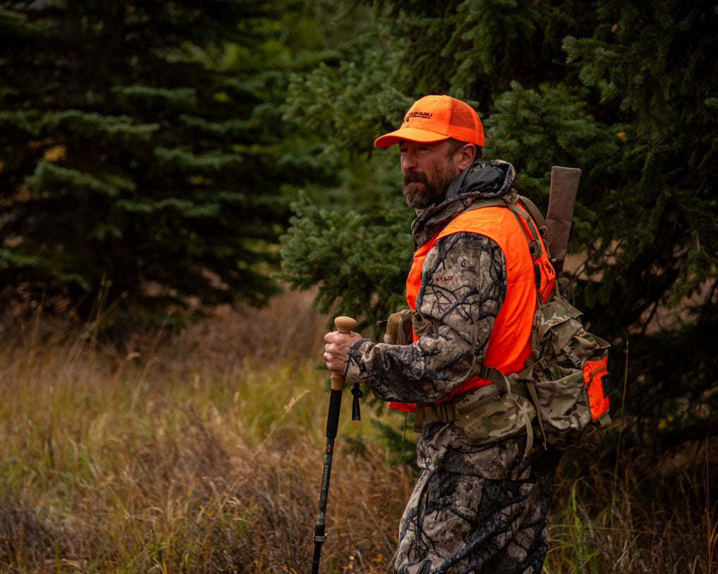 Hunter wearing Blaze Orange and camo using a cork handled trekking pole while hiking at tree&#39;s edge in a Rocky Mountain field